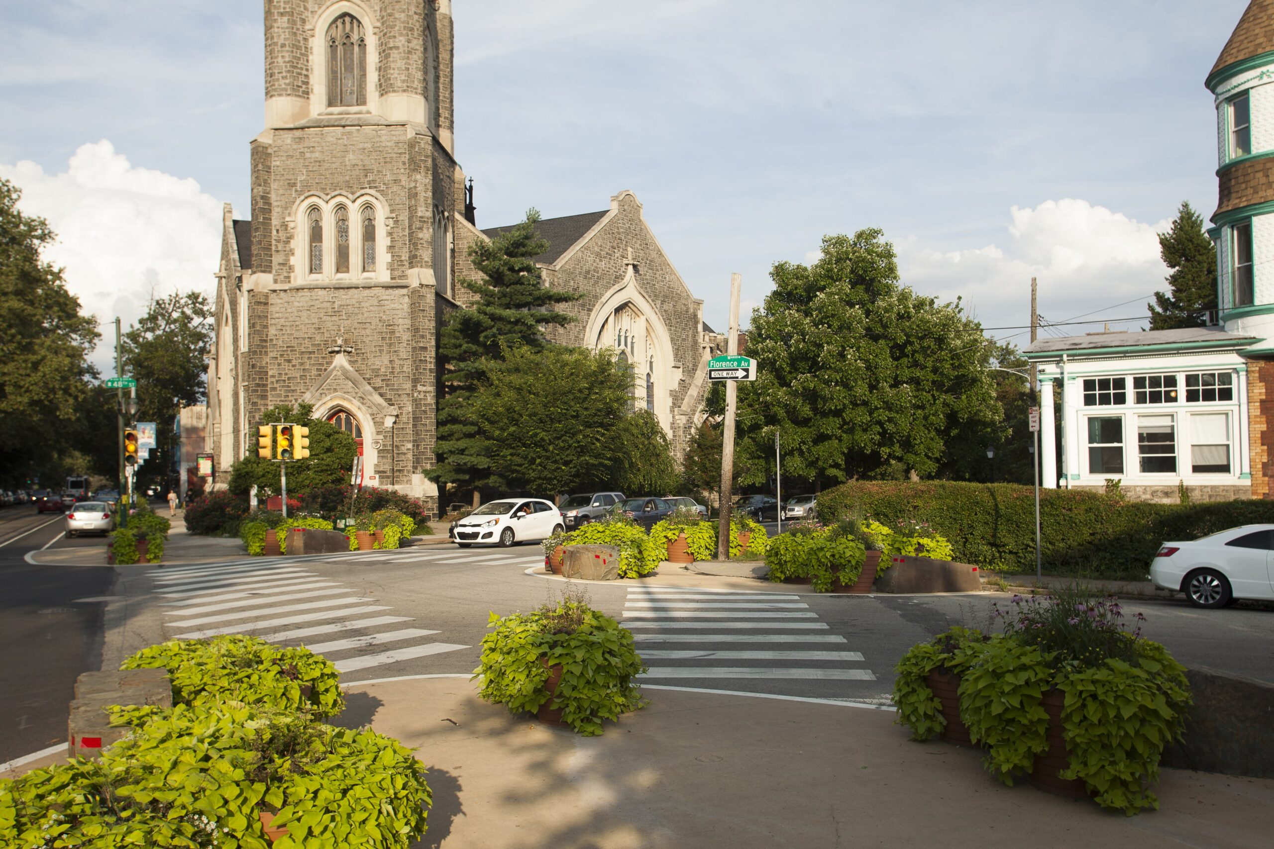 A crosswalk at Baltimore Crossing in Philadelphia, with planters filled with greenery, a historic stone church, and cars along the street.