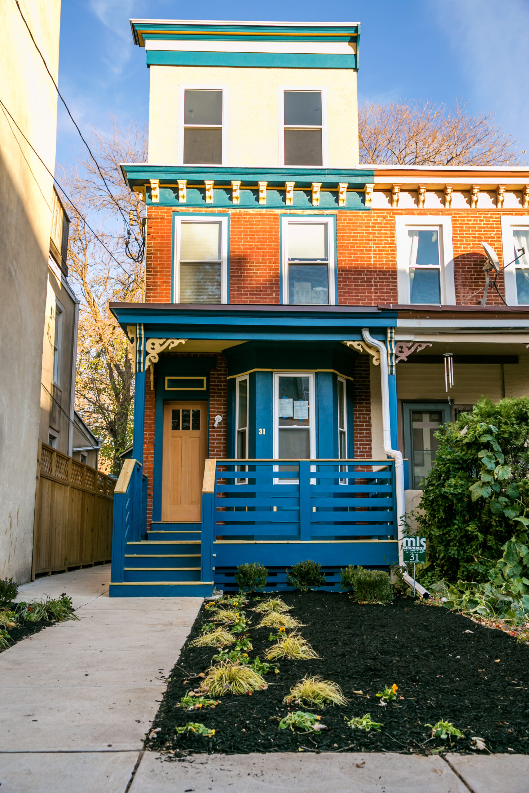 A completed renovation of a rowhouse on Preston Street with a blue exterior, landscaped yard, and wooden porch.