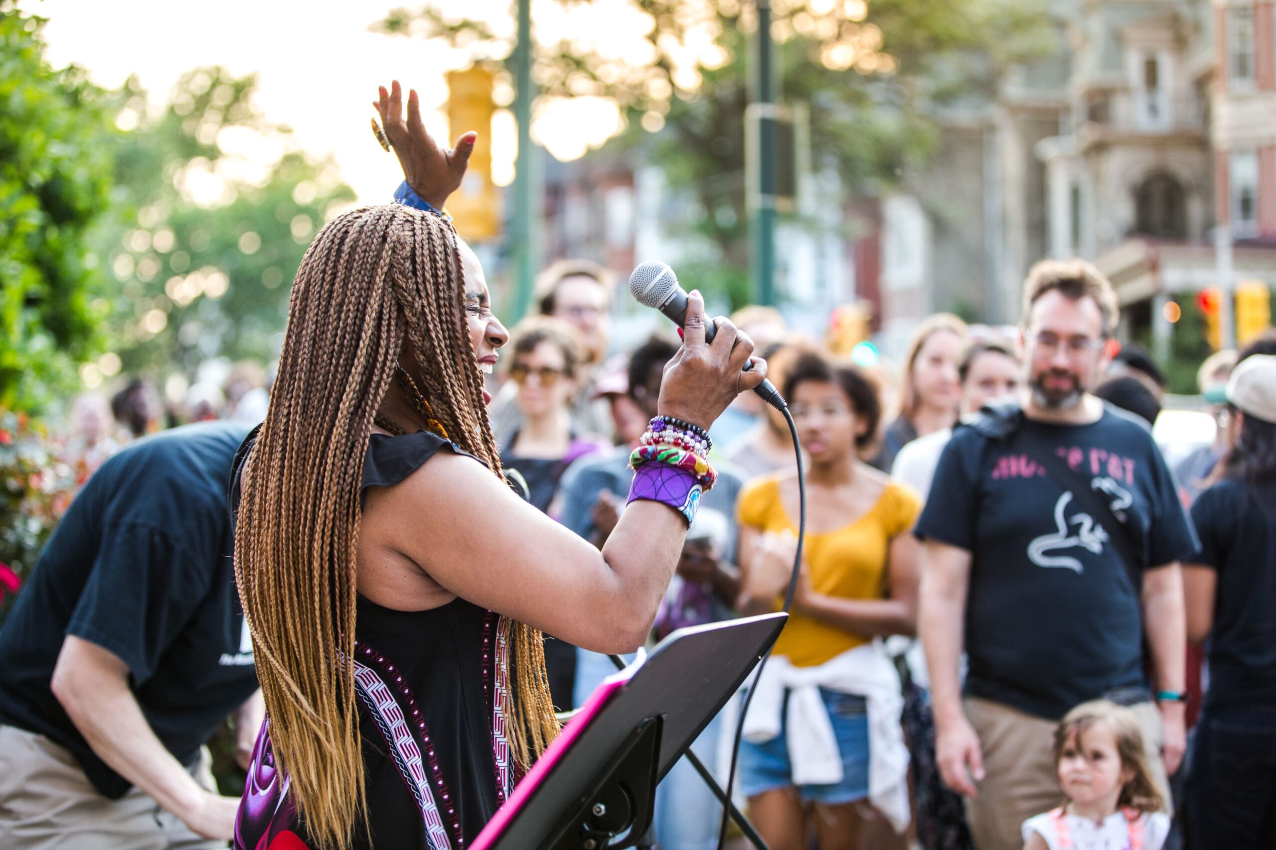 A singer with long braids performs with a microphone in front of a lively crowd at the Baltimore Avenue Dollar Stroll.