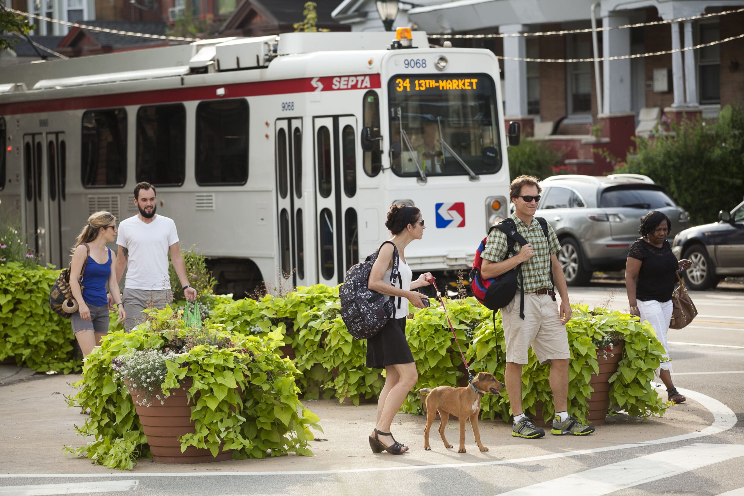 Pedestrians walking alongside a SEPTA Route 34 trolley near Baltimore Crossing, with vibrant potted plants and urban landscaping in the foreground.