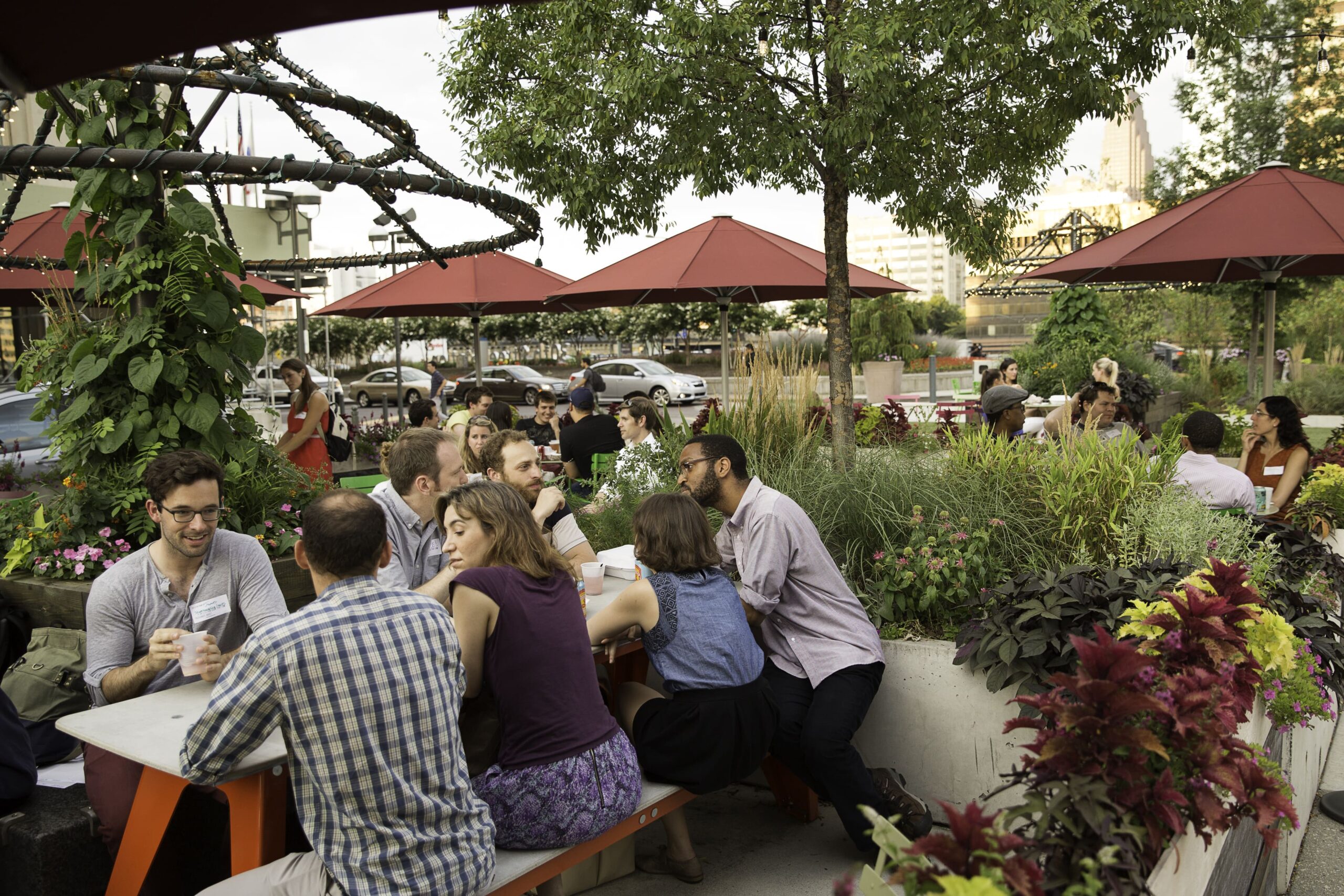 Groups of people sit at picnic tables under red umbrellas at The Porch at 30th Street Station, surrounded by lush greenery and a lively atmosphere.