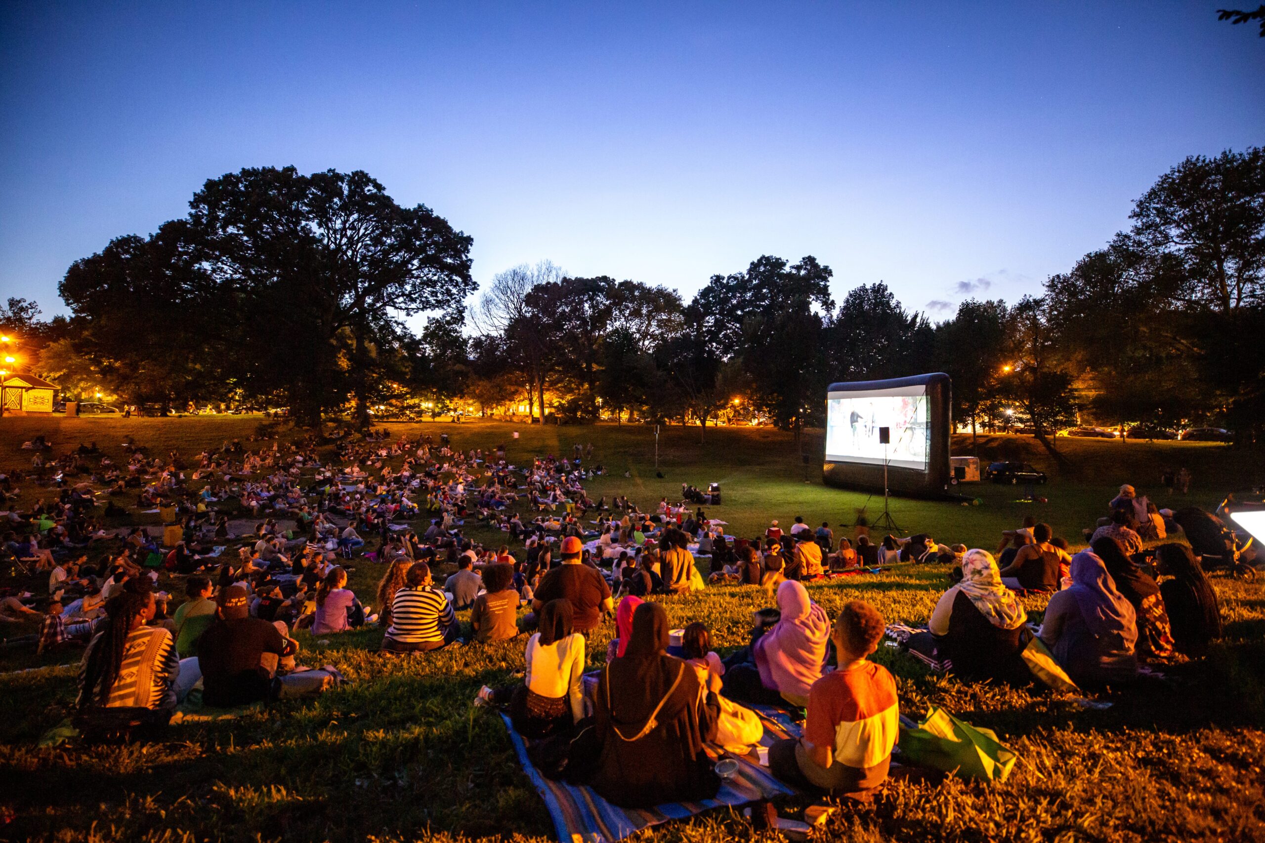 A crowd sits on the grass at Clark Park, watching an outdoor movie under the evening sky surrounded by trees and glowing lights.