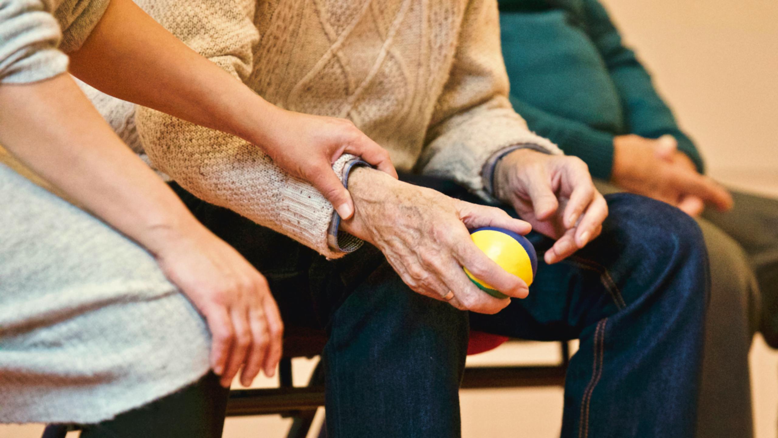 A caregiver gently holds the wrist of an elderly person holding a stress ball, symbolizing compassion and support for the elderly.