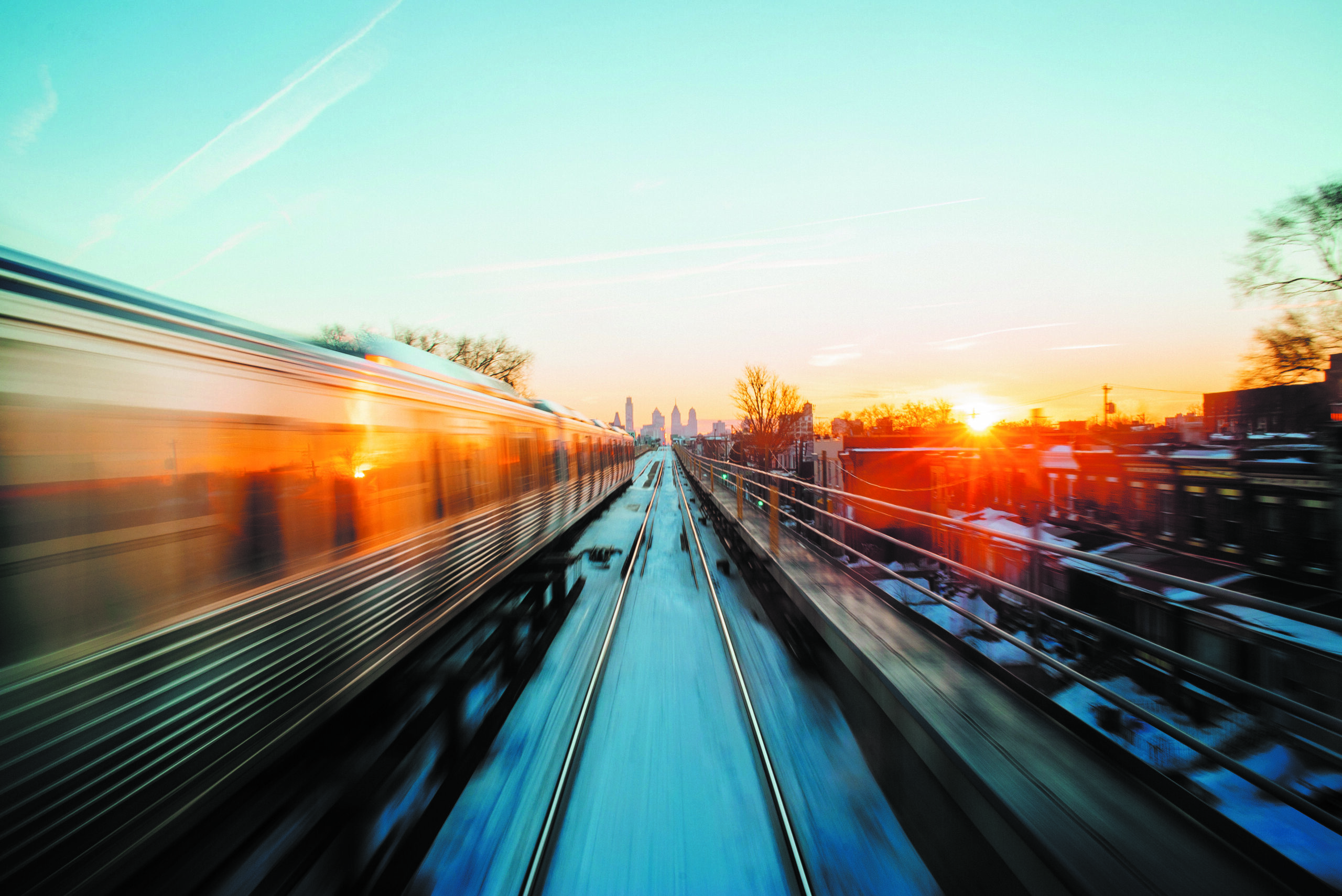 A train speeding on elevated tracks toward the Philadelphia skyline at sunset.