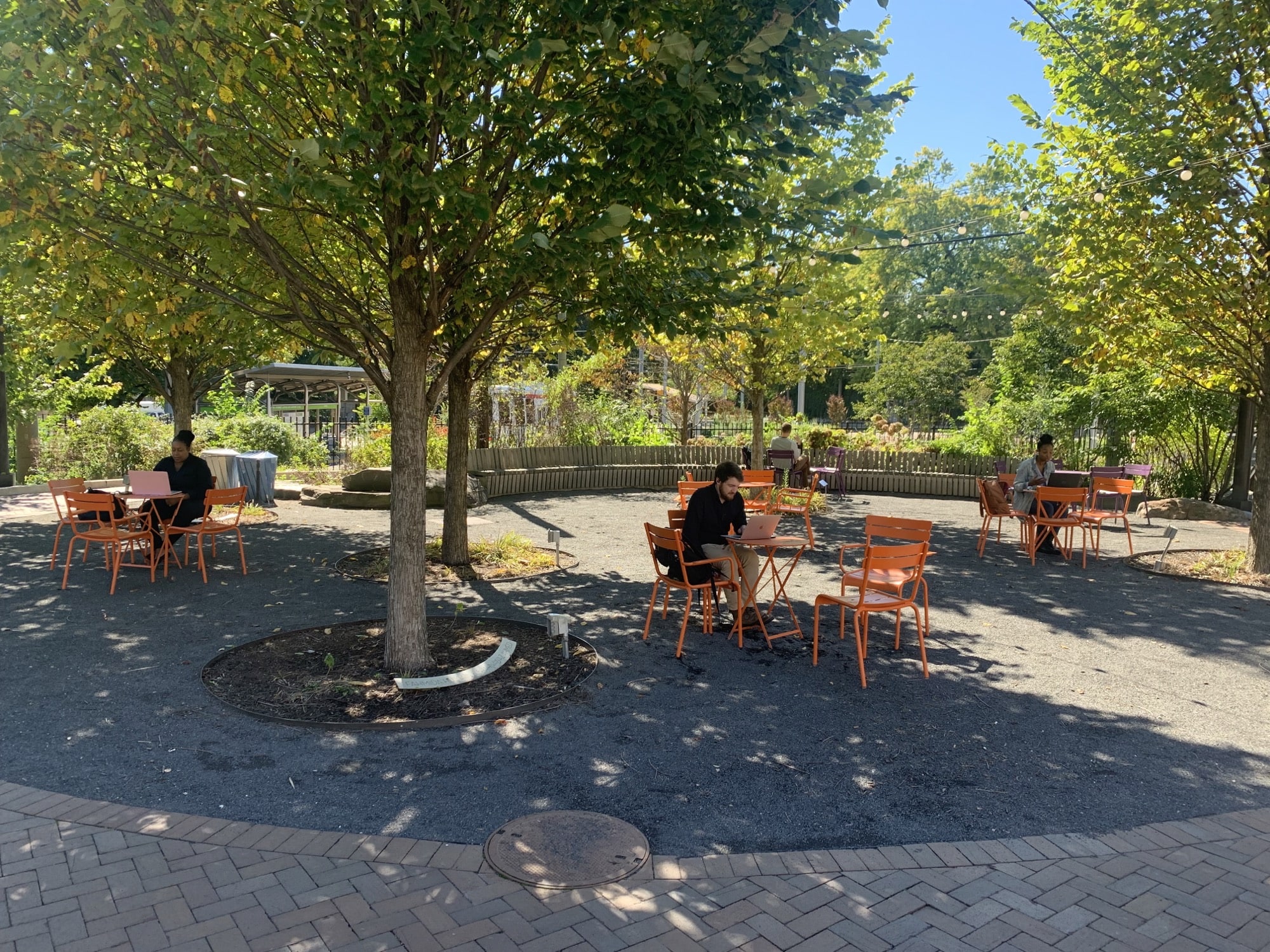 People sit at orange tables under the shade of trees at Trolley Portal Gardens, working on laptops in a calm outdoor setting.