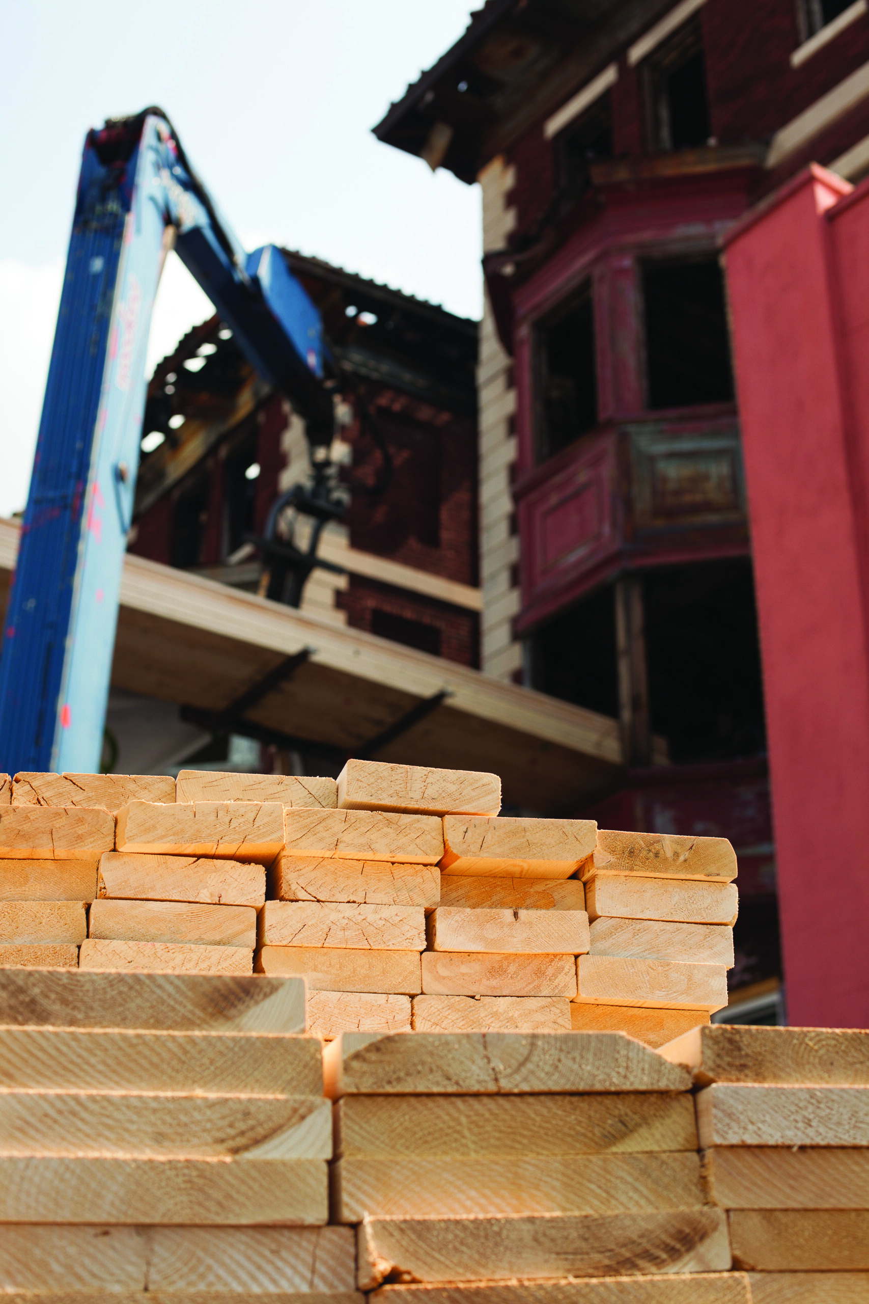 Stack of lumber with a partially demolished building and construction machinery in the background.