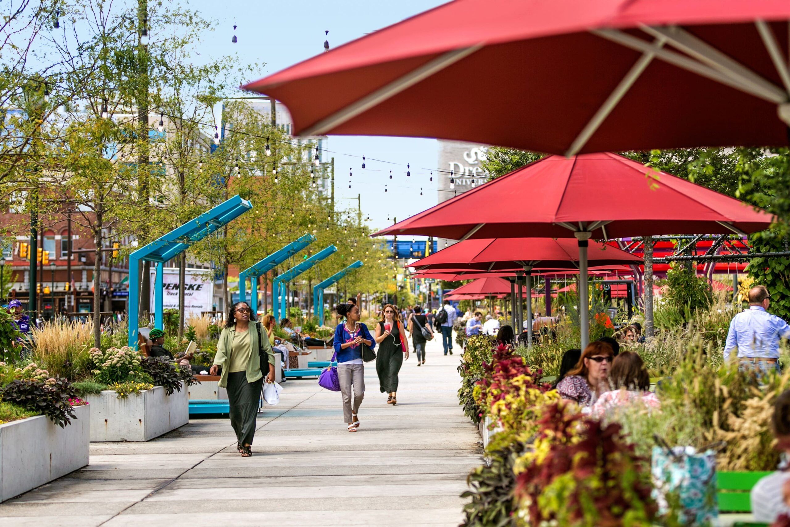 People walking and relaxing under red umbrellas at The Porch at 30th Street Station, surrounded by greenery and urban landscaping.