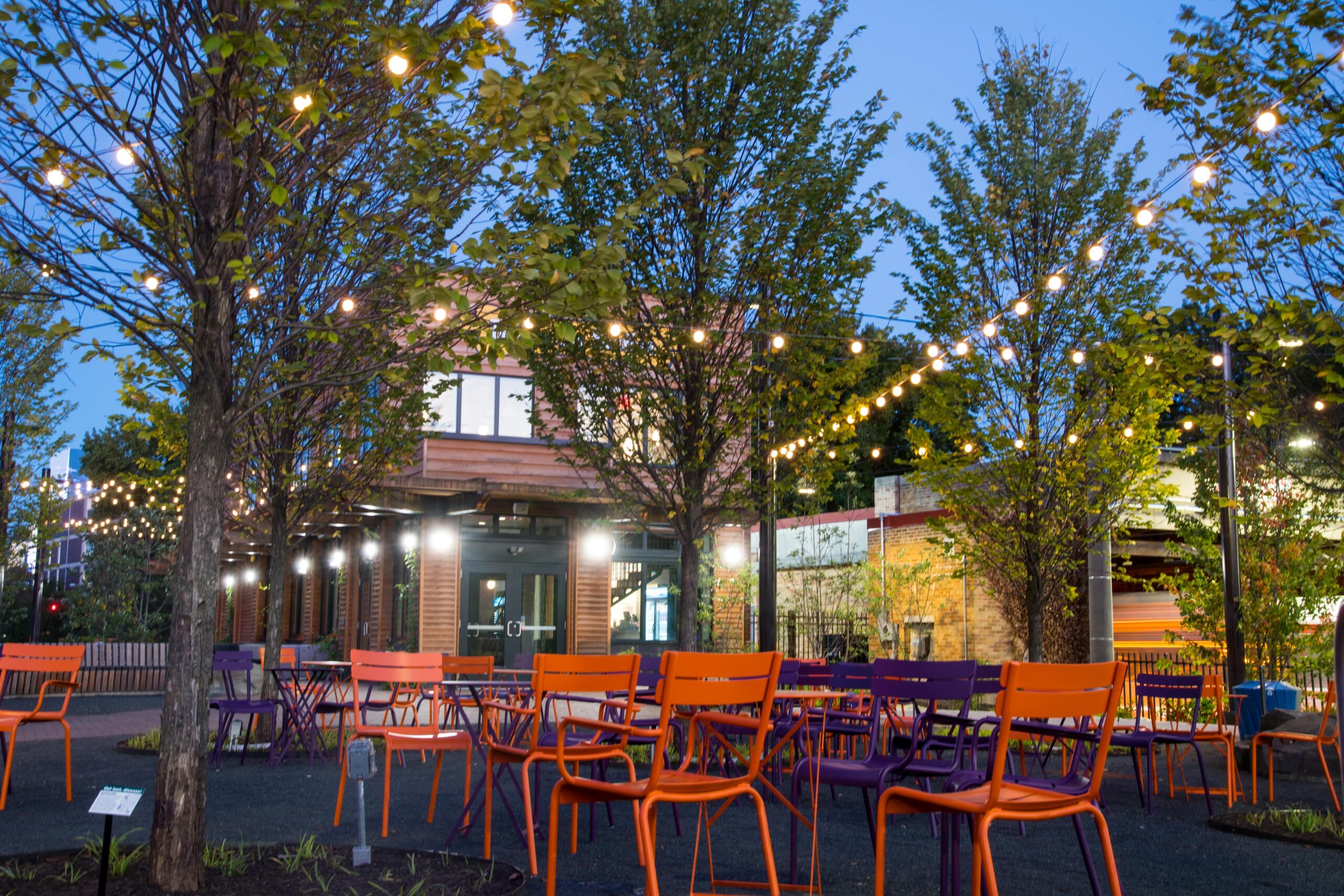 An evening view of Trolley Portal Gardens with colorful chairs, string lights, and trees creating a welcoming atmosphere.