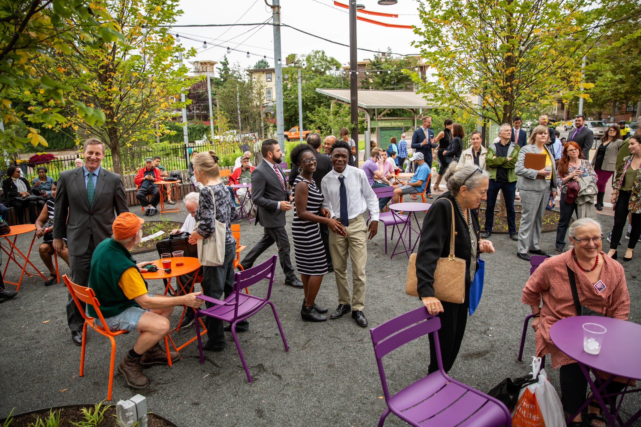 A bustling community event at the Trolley Portal Gardens opening, featuring people gathered at colorful tables and chairs, socializing and celebrating.