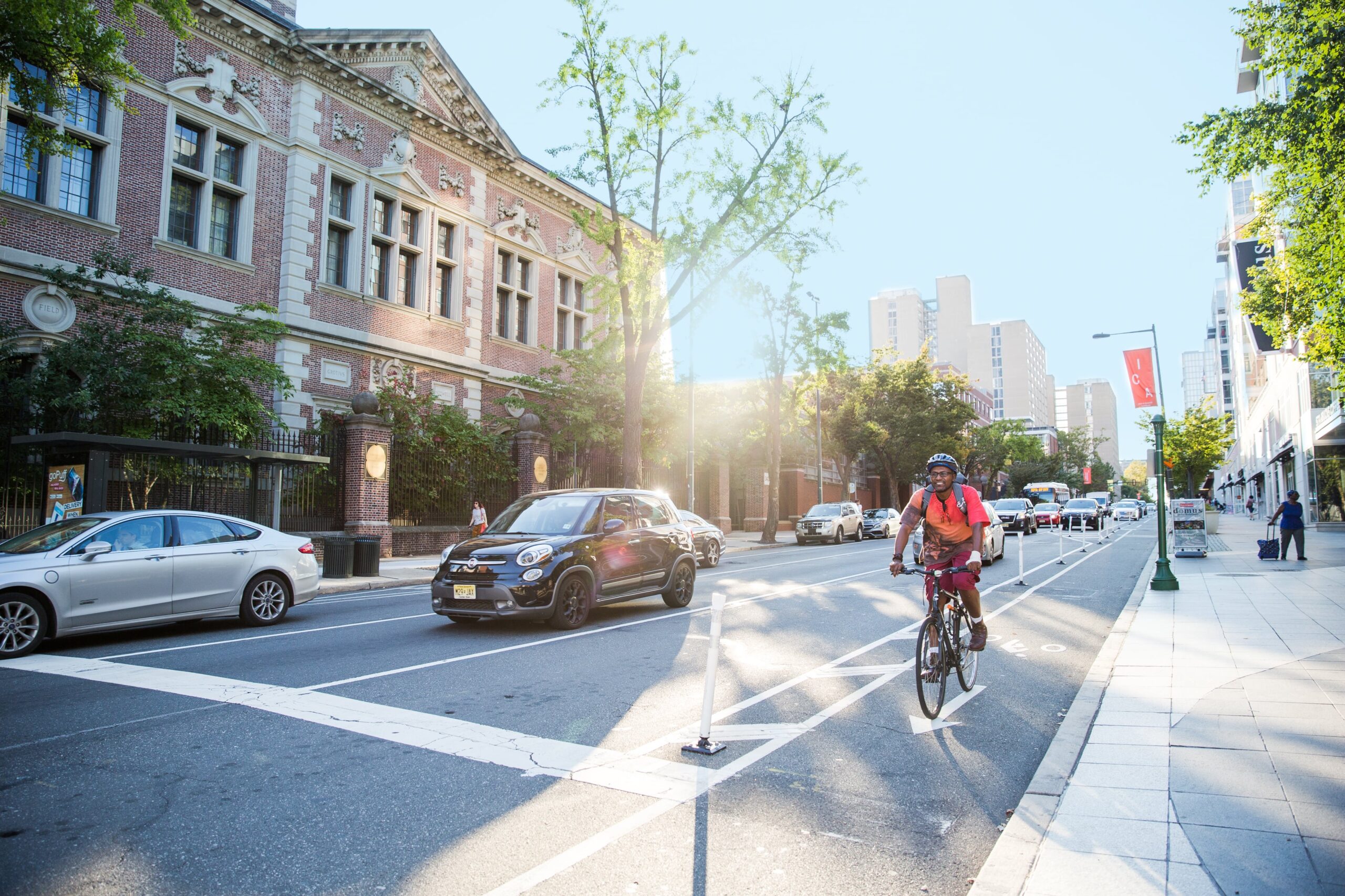 Cyclist using a protected bike lane in University City, with a sunlit street, historic architecture, and cars in the background.