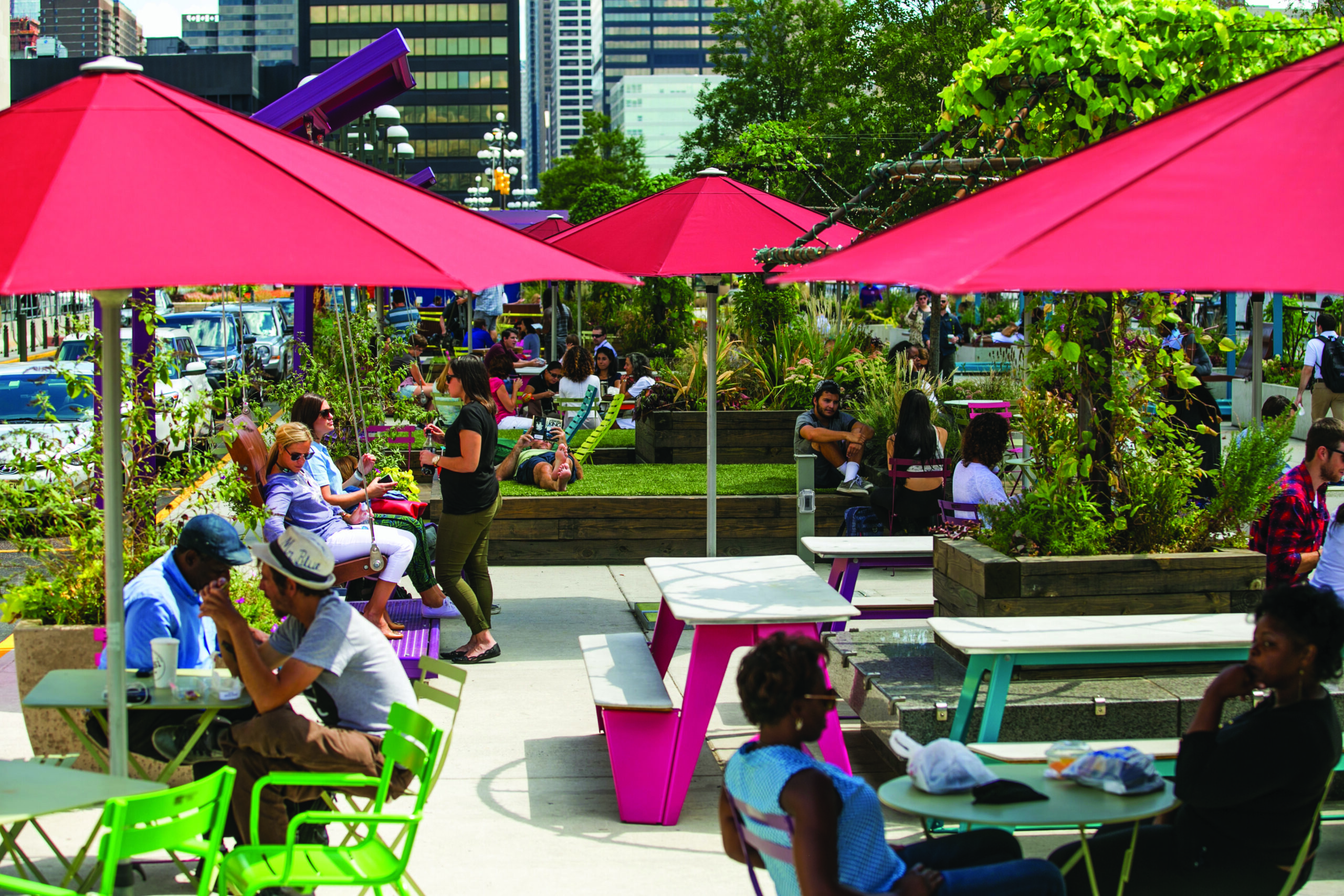 People relax on swings and enjoy outdoor seating under red umbrellas at The Porch at 30th Street Station, surrounded by lush greenery.