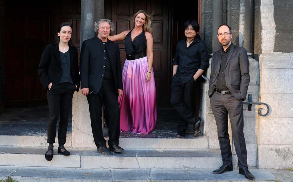 Photograph of five people standing on a staircase. Celine Scheen stands in the center, and wears a black top and a pink ombre skirt. The others stand on either side of her, dressed in all black.