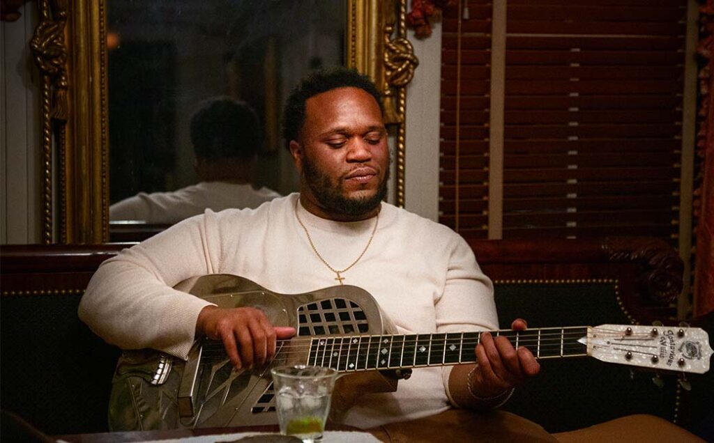 Close-up photo of singer Jontavious Willis playing a silver guitar in a dark, moody bar. He gazes at the guitar with a calm expression on his face as he plays.