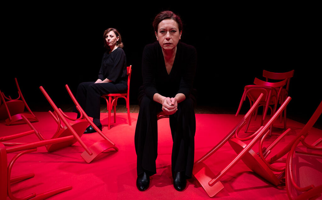 Two women sit on red chairs on a stage with a bright red floor. The woman in the center faces the camera, while the woman in the back sits at a 45-degree angle. Overturned red chairs are scattered around them.