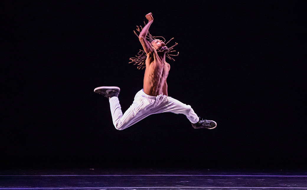 Male dancer leaps through the air on stage, one arm raised. He has long dreadlocks and wears white pants and basketball shoes