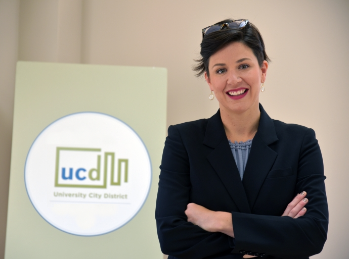 Sarah Steltz stands with her arms crossed, smiling in front of a board displaying the University City logo. She wears a black blazer.