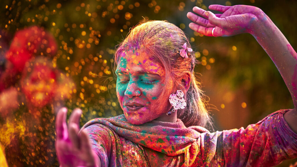 Photograph of a woman celebrating The Hindu tradition of Holi, she dances as she's covered in brightly colored powders. 