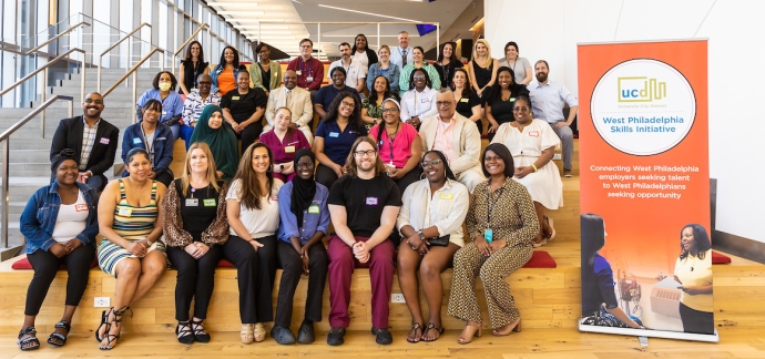 A large group of people from the Hummingbird Foundation sit on a set of stairs, smiling at the camera. To the right is a large , orange poster board promoting The Skills Initiative